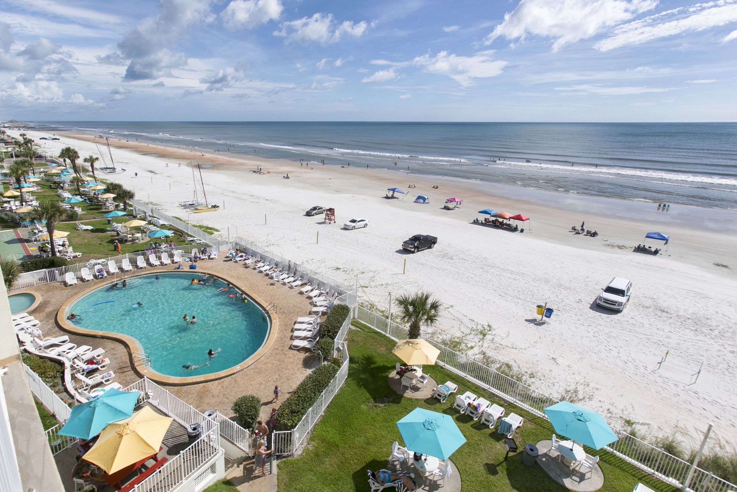 An aerial view of a beachfront resort with a pool, lounge chairs, and palm trees.